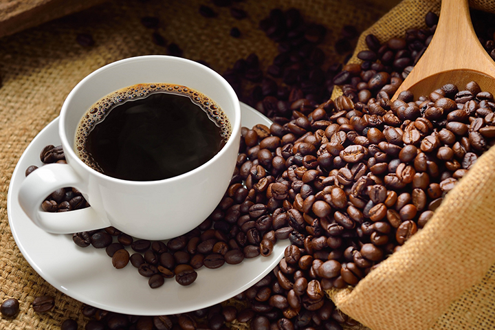 A pile of coffee beans, spilling out from a wooden scoop, with a white cup and saucer full of coffee sitting on top.